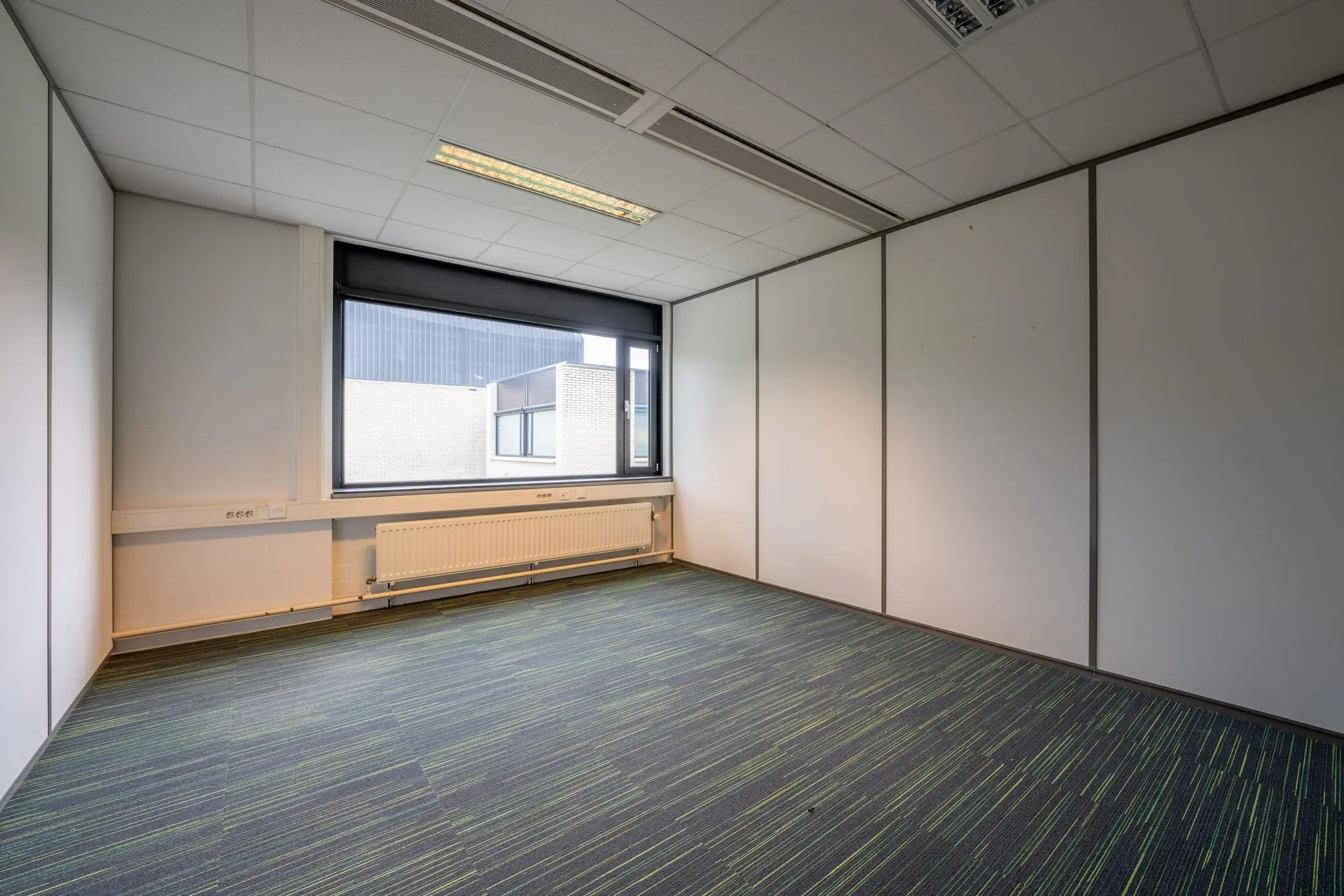 Leeg kantoor met systeemplafond, grote raam en blauwgroen gestreepte tapijttegels aan de Burgemeester Verderlaan.